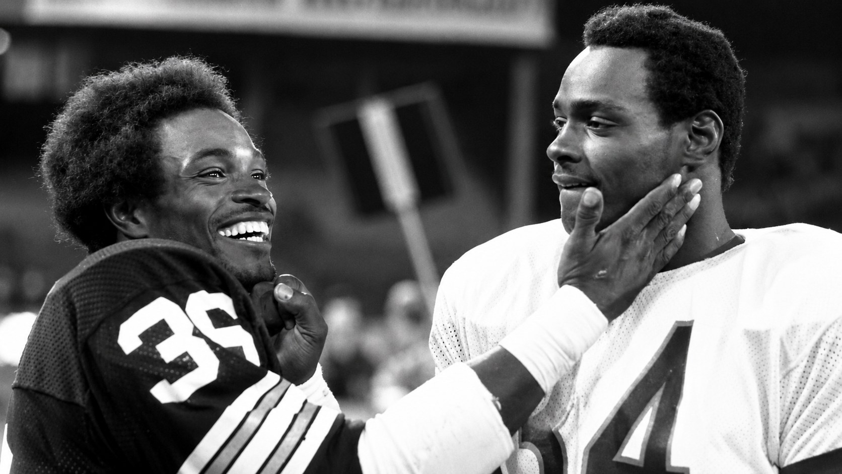 Eddie Payton, in a dark number 39 jersey, laughing with his brother Walter Payton in the Chicago Bears' white number 34 road jersey on an NFL sideline.